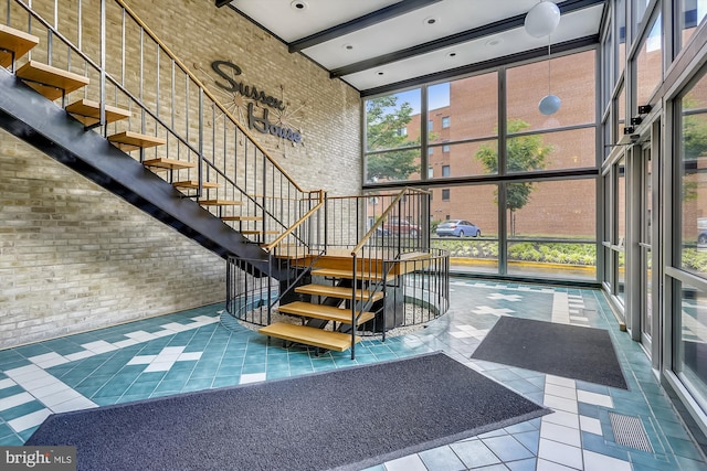 stairs with brick wall, tile patterned flooring, a towering ceiling, a wall of windows, and beamed ceiling