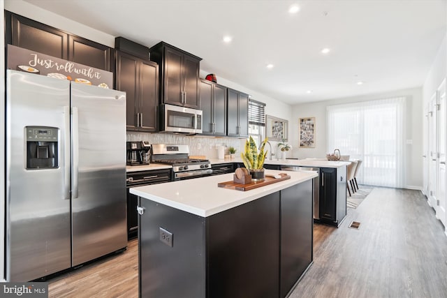 kitchen with appliances with stainless steel finishes, dark brown cabinetry, a kitchen island, decorative backsplash, and light wood-type flooring