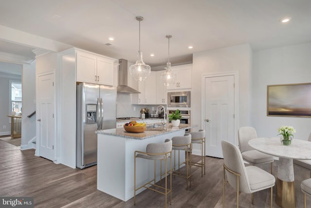 kitchen featuring a kitchen island with sink, wall chimney range hood, appliances with stainless steel finishes, decorative light fixtures, and white cabinetry