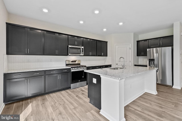 kitchen with a kitchen island with sink, sink, appliances with stainless steel finishes, light hardwood / wood-style floors, and light stone counters