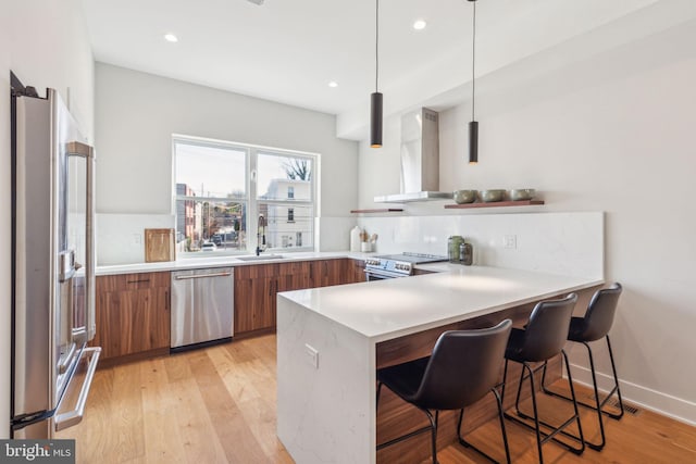 kitchen with kitchen peninsula, stainless steel appliances, sink, wall chimney range hood, and hanging light fixtures