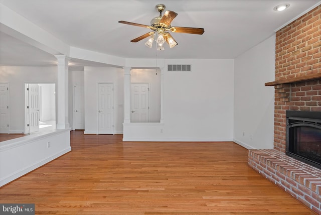 unfurnished living room with a fireplace, visible vents, a ceiling fan, light wood finished floors, and decorative columns