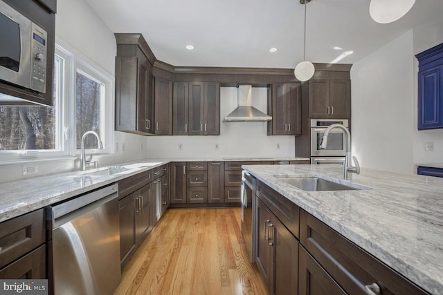 kitchen with stainless steel appliances, sink, light stone counters, wall chimney exhaust hood, and hanging light fixtures