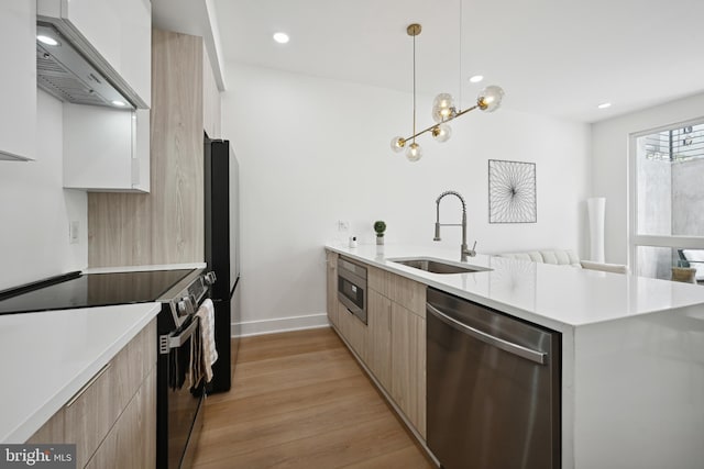 kitchen with appliances with stainless steel finishes, wall chimney range hood, sink, light brown cabinets, and hanging light fixtures