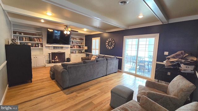 living room featuring a fireplace, built in shelves, ceiling fan, light hardwood / wood-style flooring, and beamed ceiling