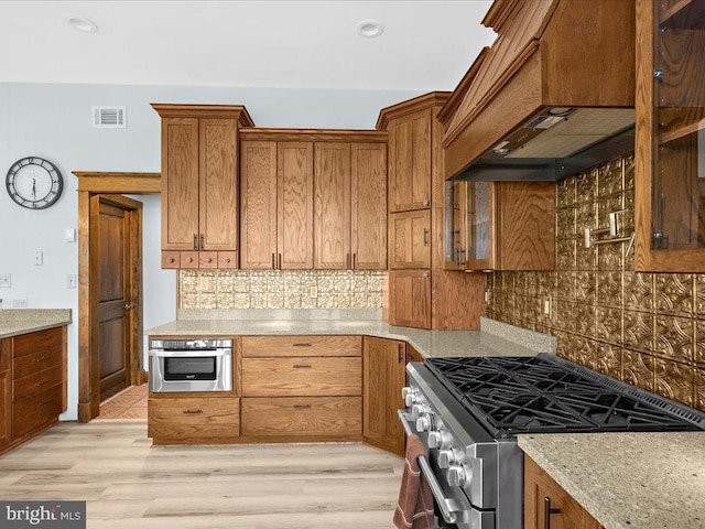 kitchen featuring decorative backsplash, appliances with stainless steel finishes, light stone countertops, premium range hood, and light hardwood / wood-style floors