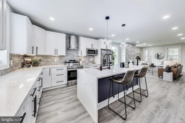 kitchen with appliances with stainless steel finishes, wall chimney exhaust hood, pendant lighting, a center island with sink, and white cabinets