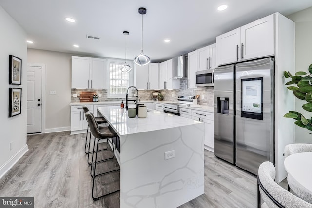 kitchen featuring wall chimney exhaust hood, an island with sink, pendant lighting, white cabinets, and appliances with stainless steel finishes