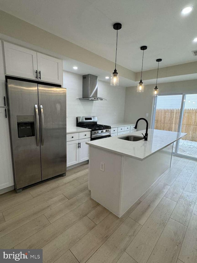 kitchen featuring white cabinets, wall chimney range hood, stainless steel appliances, sink, and a kitchen island with sink