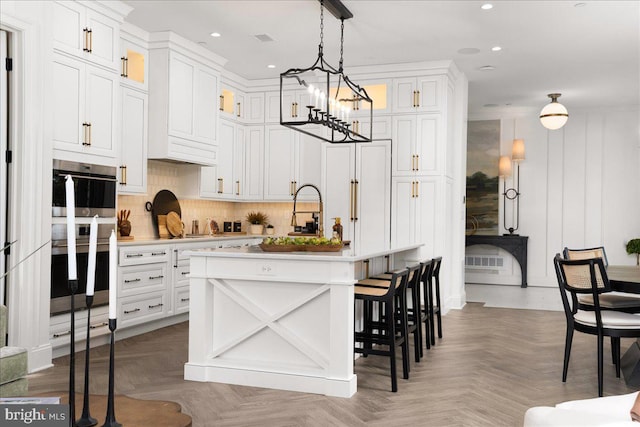 kitchen with parquet flooring, hanging light fixtures, an island with sink, double oven, and white cabinets