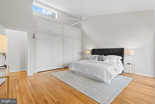 bedroom with two closets, baseboards, light wood-style flooring, a towering ceiling, and a ceiling fan