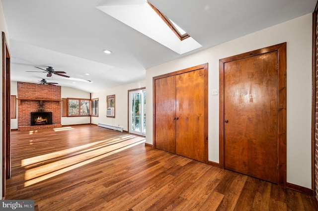 unfurnished living room with a brick fireplace, vaulted ceiling with skylight, ceiling fan, baseboard heating, and dark wood-type flooring