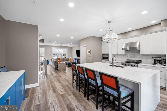 kitchen featuring sink, white cabinetry, an island with sink, ceiling fan with notable chandelier, and wall chimney range hood