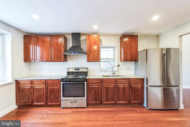 kitchen featuring wall chimney range hood, sink, hardwood / wood-style flooring, light stone countertops, and stainless steel appliances