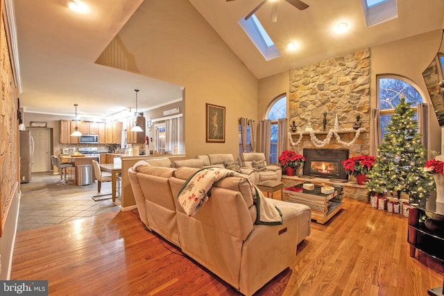 living room featuring high vaulted ceiling, a skylight, light hardwood / wood-style flooring, ceiling fan, and a fireplace