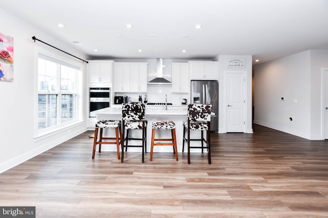 kitchen featuring white cabinetry, wall chimney exhaust hood, stainless steel appliances, a breakfast bar area, and a kitchen island with sink