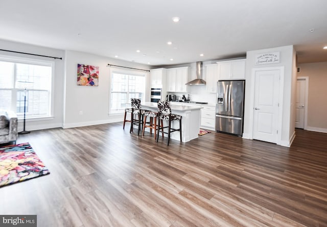 kitchen featuring a kitchen bar, appliances with stainless steel finishes, a kitchen island with sink, wall chimney range hood, and white cabinetry