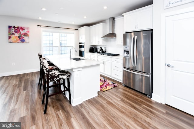 kitchen with white cabinetry, wall chimney exhaust hood, a kitchen breakfast bar, an island with sink, and appliances with stainless steel finishes