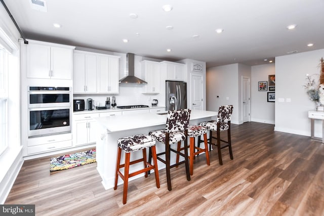 kitchen with appliances with stainless steel finishes, wall chimney exhaust hood, a breakfast bar, white cabinets, and an island with sink