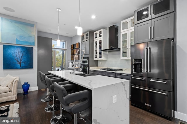 kitchen with appliances with stainless steel finishes, backsplash, a kitchen island with sink, wall chimney range hood, and decorative light fixtures