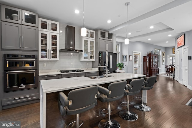 kitchen with gray cabinetry, wall chimney exhaust hood, hanging light fixtures, high end black fridge, and a center island with sink