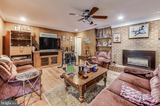 living room with ceiling fan, a brick fireplace, wood walls, light hardwood / wood-style floors, and crown molding