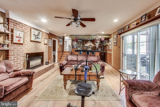 living room featuring wood walls, a brick fireplace, ceiling fan, light wood-type flooring, and crown molding