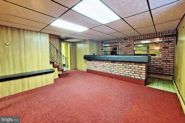 basement featuring a drop ceiling, wood walls, indoor bar, a baseboard radiator, and carpet floors