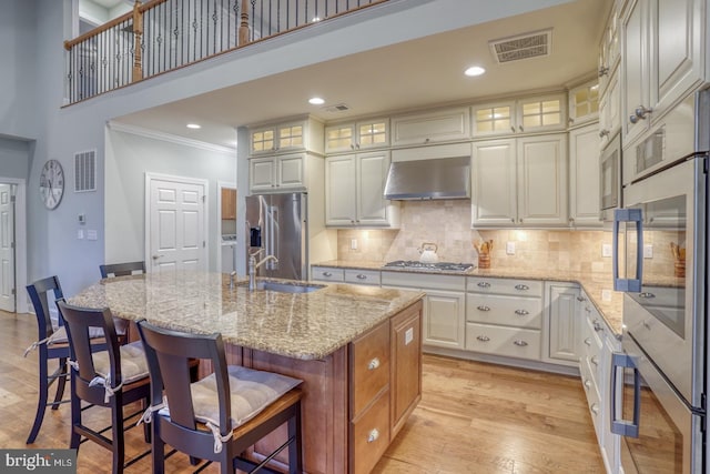 kitchen with wall chimney range hood, sink, a kitchen island with sink, stainless steel appliances, and light stone counters