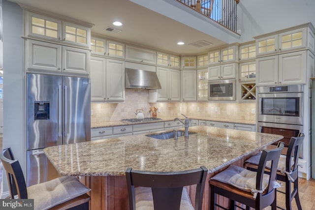 kitchen featuring appliances with stainless steel finishes, sink, a center island with sink, and wall chimney range hood