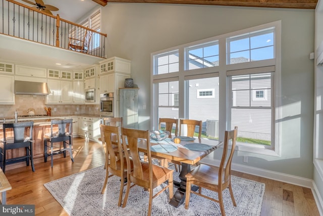 dining space with ceiling fan, beam ceiling, light wood-type flooring, and a high ceiling