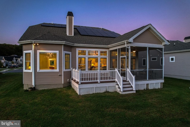 back house at dusk with a wooden deck, solar panels, and a yard