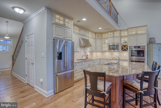 kitchen featuring stainless steel appliances, a kitchen island with sink, pendant lighting, wall chimney exhaust hood, and sink