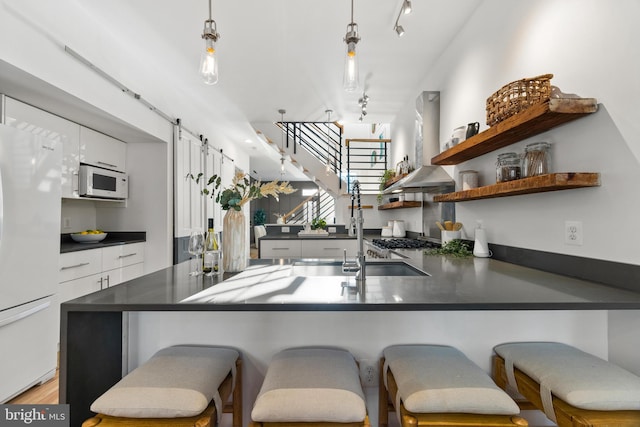 kitchen featuring white cabinetry, wall chimney exhaust hood, white appliances, a kitchen bar, and hanging light fixtures