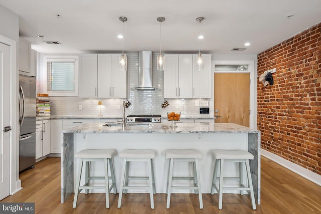 kitchen with hanging light fixtures, freestanding refrigerator, white cabinets, wall chimney range hood, and an island with sink