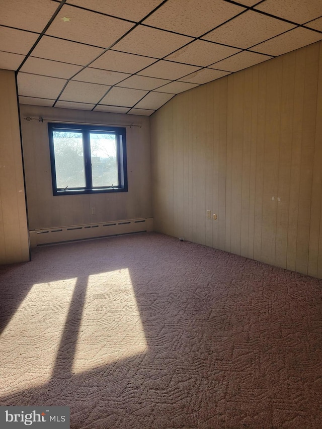 carpeted empty room featuring a paneled ceiling, wood walls, and a baseboard heating unit