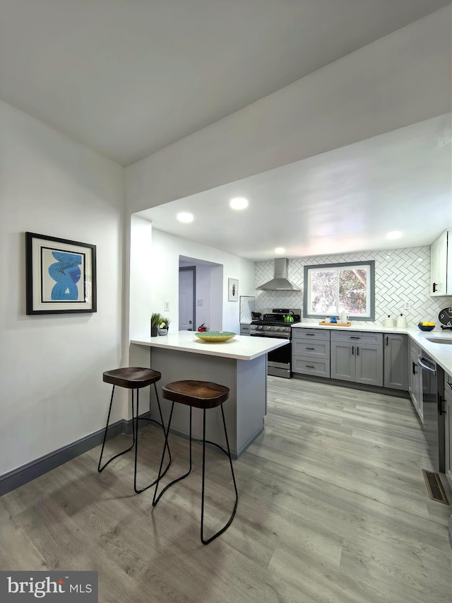 kitchen featuring stove, wall chimney exhaust hood, gray cabinets, kitchen peninsula, and a breakfast bar area