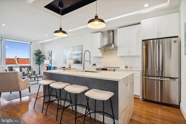 kitchen with wall chimney exhaust hood, sink, a center island with sink, white cabinetry, and stainless steel refrigerator