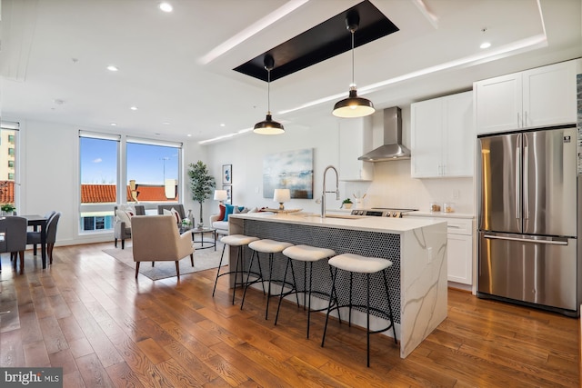 kitchen with a kitchen island with sink, wall chimney range hood, decorative light fixtures, white cabinetry, and stainless steel refrigerator