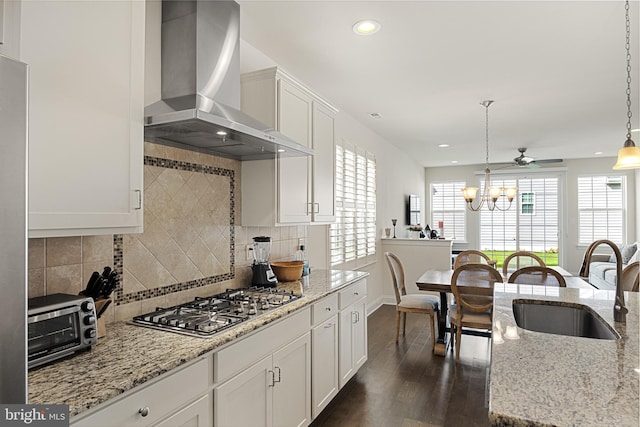 kitchen with light stone counters, wall chimney exhaust hood, stainless steel gas cooktop, white cabinets, and hanging light fixtures