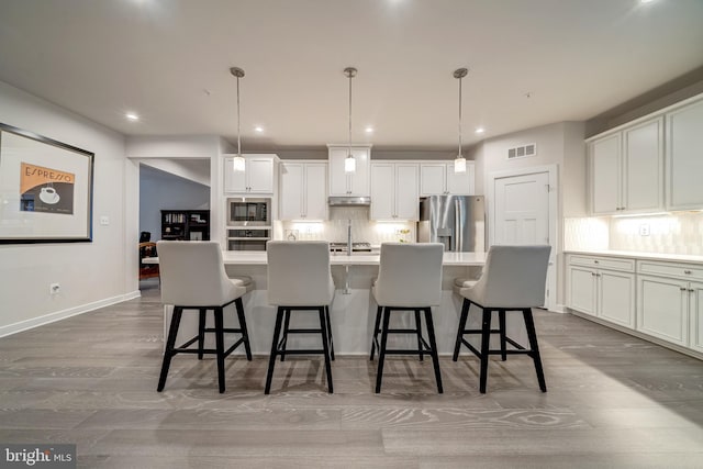 kitchen featuring appliances with stainless steel finishes, a kitchen island with sink, hanging light fixtures, white cabinetry, and a kitchen bar