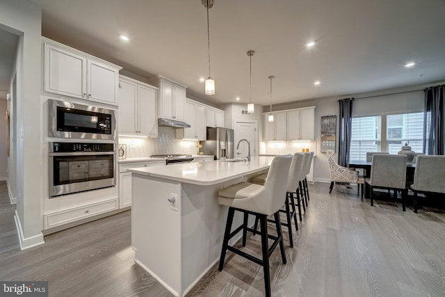 kitchen featuring tasteful backsplash, decorative light fixtures, a center island with sink, stainless steel appliances, and white cabinets