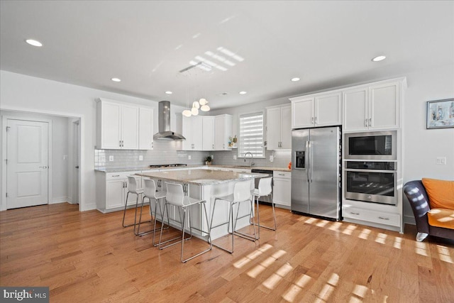 kitchen with wall chimney exhaust hood, a center island, a breakfast bar area, appliances with stainless steel finishes, and white cabinetry