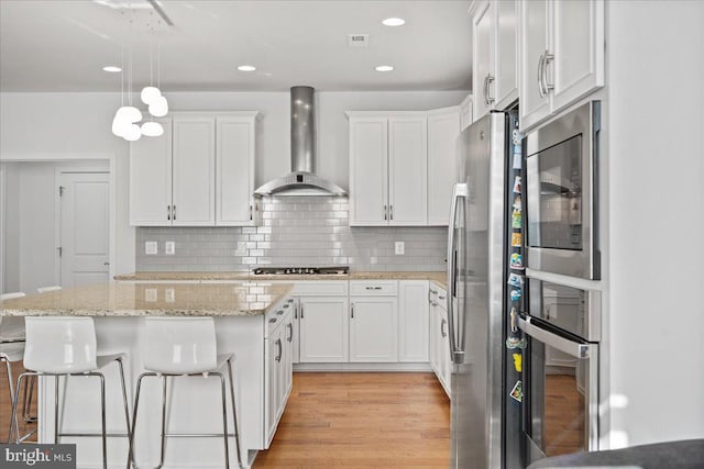 kitchen with stainless steel appliances, decorative light fixtures, wall chimney range hood, and white cabinetry