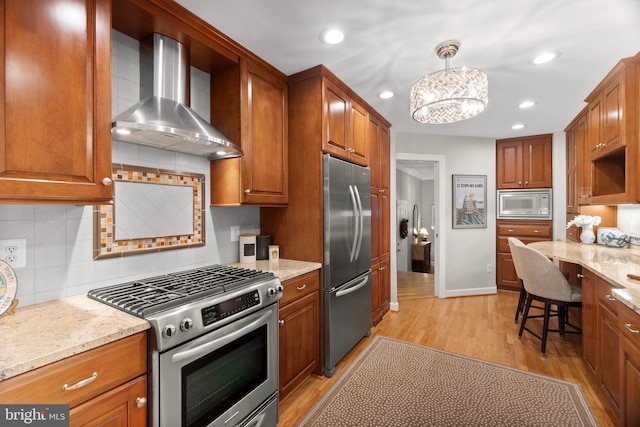 kitchen featuring wall chimney range hood, hanging light fixtures, decorative backsplash, appliances with stainless steel finishes, and light stone counters