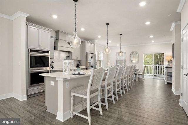 kitchen with pendant lighting, custom exhaust hood, stainless steel appliances, a kitchen breakfast bar, and a center island with sink