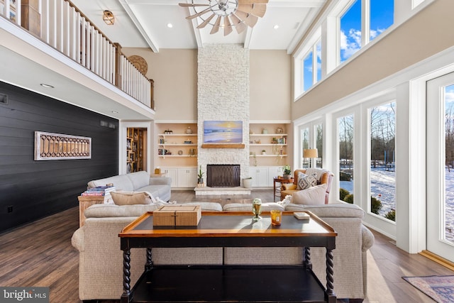 living room featuring a towering ceiling, beam ceiling, built in features, wood-type flooring, and a stone fireplace