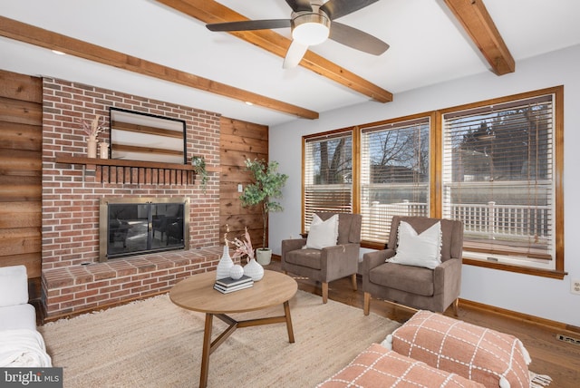 living room featuring beamed ceiling, a ceiling fan, wood finished floors, baseboards, and a brick fireplace