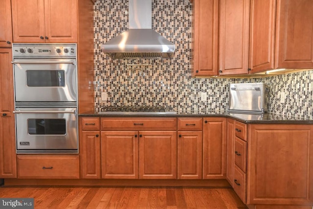 kitchen with light wood-type flooring, decorative backsplash, wall chimney exhaust hood, and appliances with stainless steel finishes