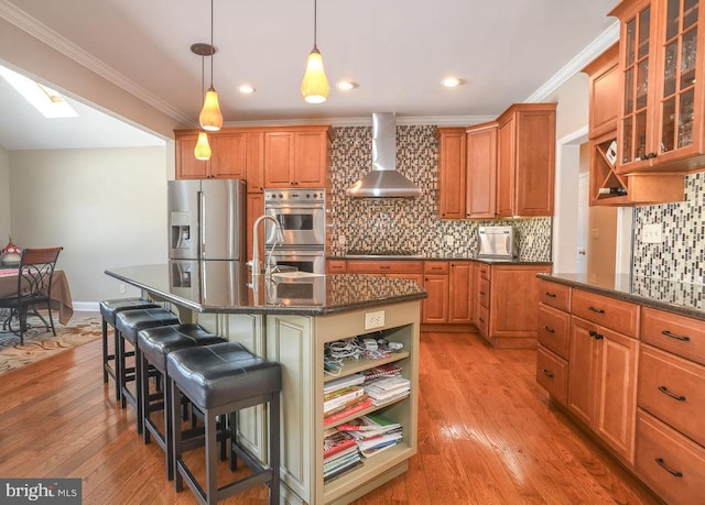 kitchen featuring wall chimney range hood, stainless steel appliances, dark stone countertops, a kitchen breakfast bar, and a center island with sink